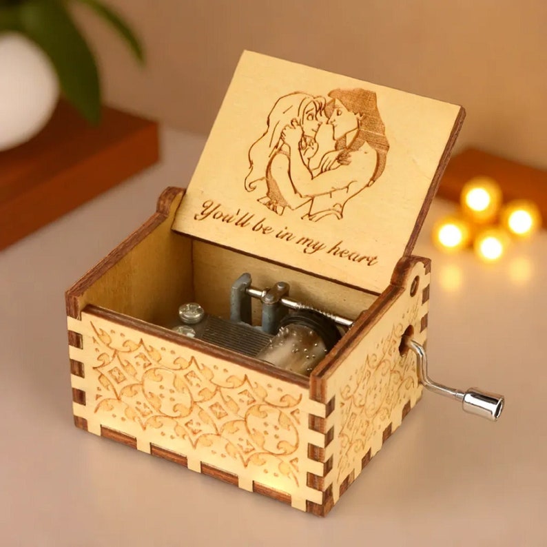 May include: A wooden music box with a hinged lid, featuring an engraved image of two figures embracing. The lid also has the text "You'll be in my heart." The box has a decorative pattern on the sides and a silver crank.