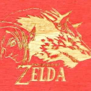 May include: A red wooden surface with a gold-colored engraved design featuring a profile of a person and a boar's head. The text "THE LEGEND OF ZELDA" is below the images.