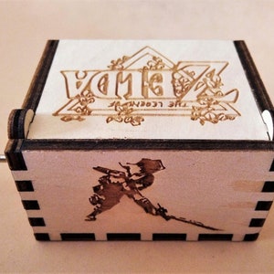 May include: Wooden music box featuring the text "ZELDA THE LEGEND OF ZELDA" and a character illustration. The box has a crank and a laser-etched design. The box is made of light-colored wood with dark brown edges.