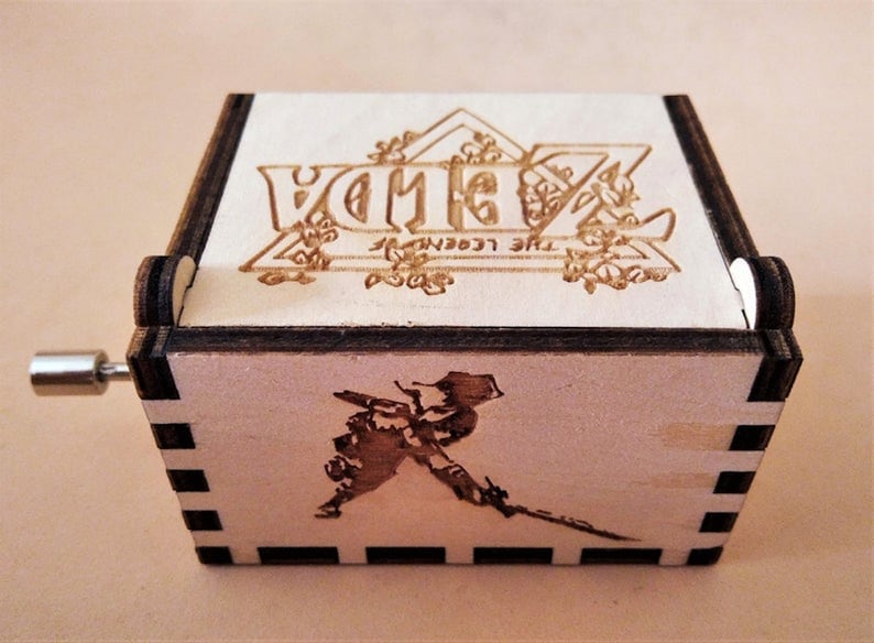 May include: Wooden music box featuring the text "ZELDA THE LEGEND OF ZELDA" and a character illustration. The box has a crank and a laser-etched design. The box is made of light-colored wood with dark brown edges.