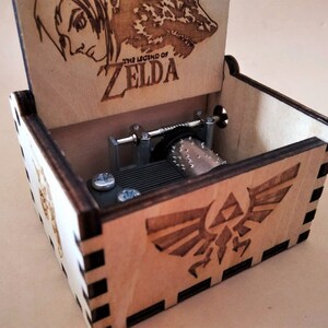 May include: A wooden music box with the words "THE LEGEND OF ZELDA" engraved on the lid. The box features a Triforce symbol and a detailed illustration. The interior mechanism is visible.
