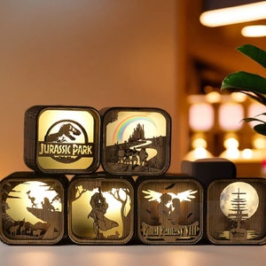May include: A collection of illuminated wooden cube lamps featuring various designs. One lamp displays the "Jurassic Park" logo, while others showcase scenes from Final Fantasy VIII and other fantasy themes. The lamps have a warm, golden glow.