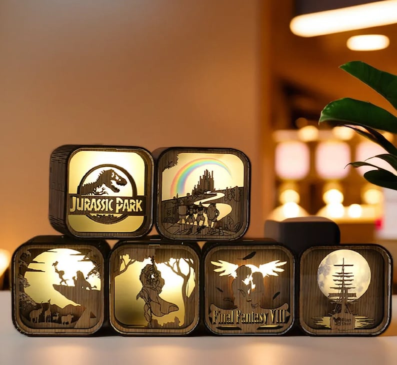 May include: A collection of illuminated wooden cube lamps featuring various designs. One lamp displays the "Jurassic Park" logo, while others showcase scenes from Final Fantasy VIII and other fantasy themes. The lamps have a warm, golden glow.