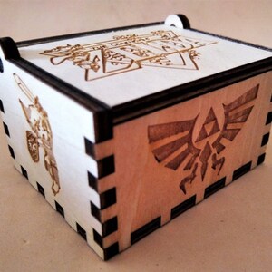 May include: A wooden box with laser-etched designs. The box features a design of a knight with a sword and shield, the Triforce symbol, and text that reads "THE LEGEND". The box is made of light-colored wood with dark edges.