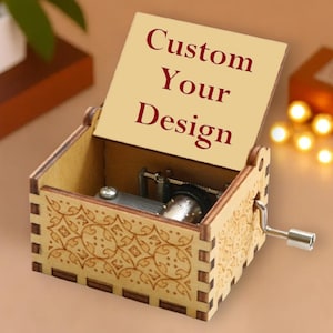 May include: A wooden music box with a hinged lid, featuring the text "Custom Your Design" in burgundy. The box has a decorative floral pattern etched on the sides and a silver crank. The interior mechanism is visible.