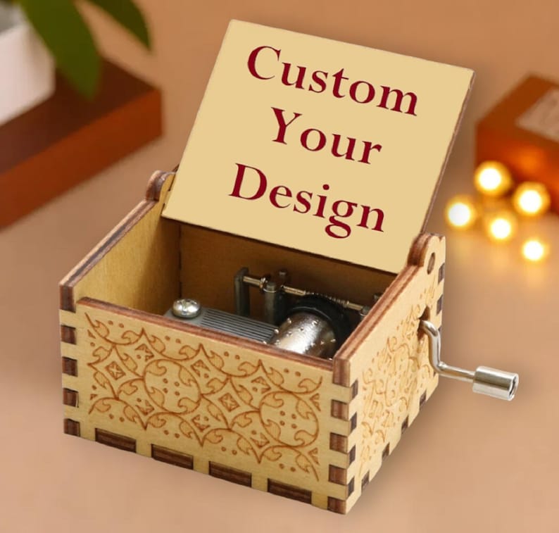 May include: A wooden music box with a hinged lid, featuring the text "Custom Your Design" in burgundy. The box has a decorative floral pattern etched on the sides and a silver crank. The interior mechanism is visible.