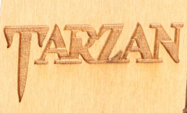 May include: Close-up of the word "TARZAN" carved into a light brown wooden surface. The letters are raised and have a textured appearance, creating a three-dimensional effect. The wood grain is visible.