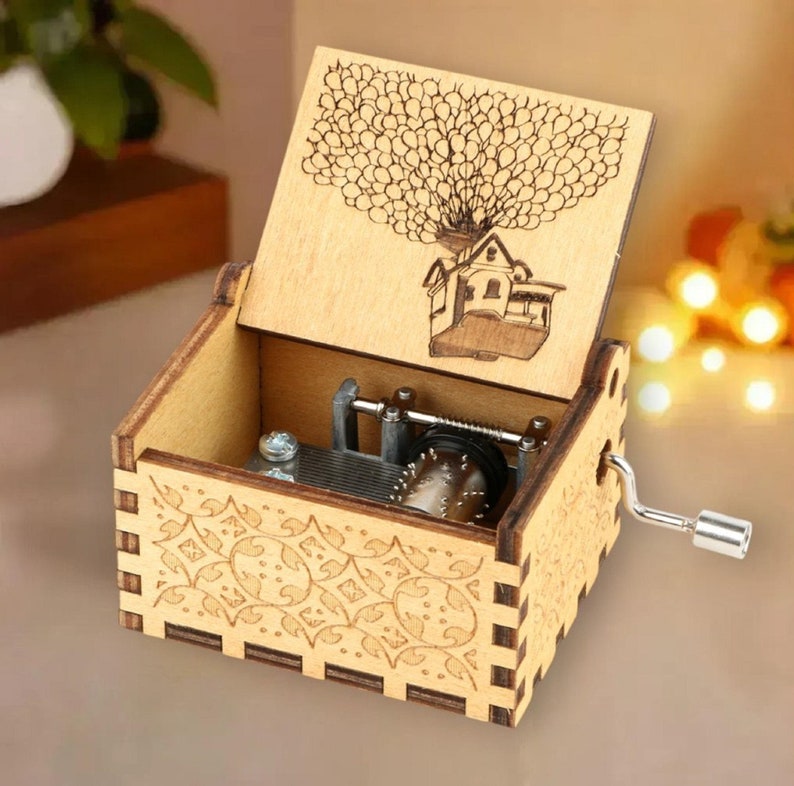 May include: A wooden music box with a hand crank and a detailed floral pattern on the sides. The lid features an etched design of a house with a tree. The music box mechanism is visible inside.