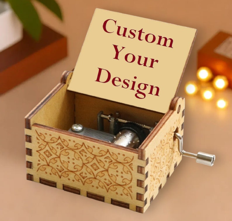 May include: A wooden music box with an open lid displaying the text "Custom Your Design" in burgundy. The box features a decorative floral pattern and a silver crank. The music box is a light brown color.