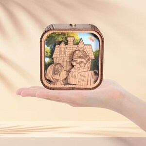 May include: A wooden music box with a carved design featuring a house and two figures kissing. The box has a light brown finish and a small, round knob on top. The words "Love You" are carved on the box.
