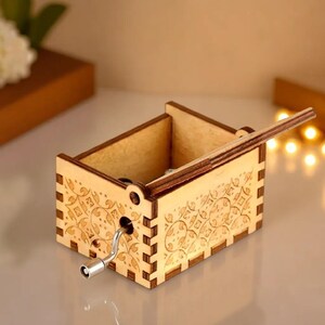 May include: A small, wooden music box with a hand crank and decorative floral patterns etched on the sides. The box is made of light-colored wood and has a hinged lid. The crank is silver-colored.