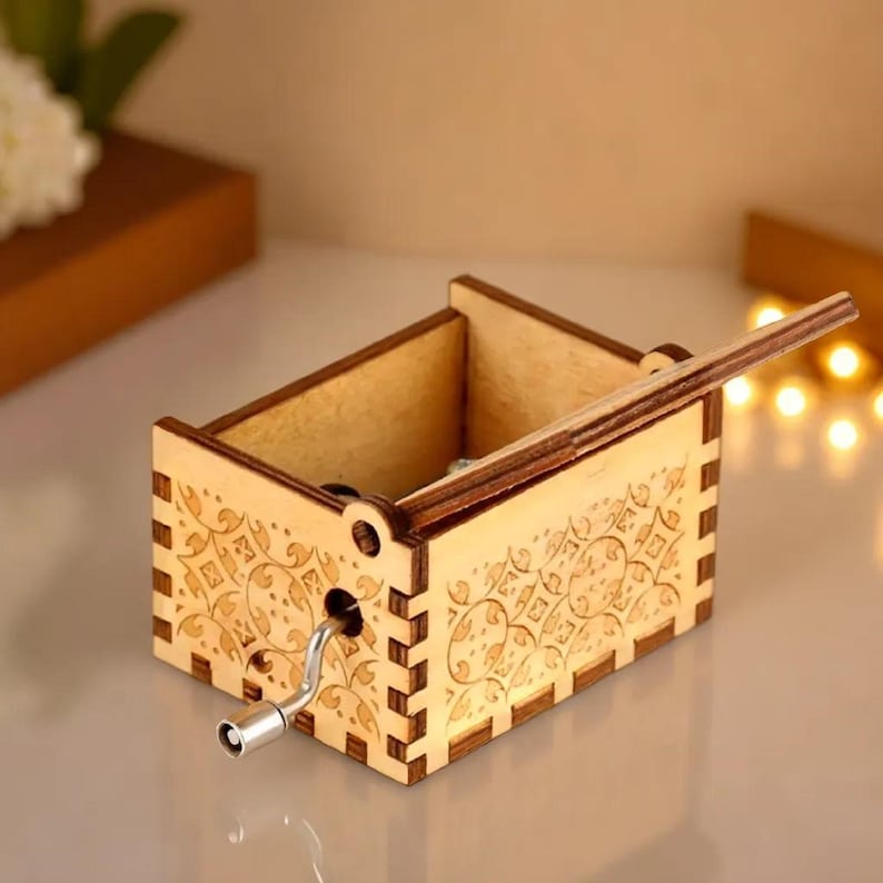 May include: A small, wooden music box with a hand crank and decorative floral patterns etched on the sides. The box is made of light-colored wood and has a hinged lid. The crank is silver-colored.