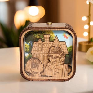 May include: A wooden music box featuring a laser-cut design of a couple and a house. The box has a square shape with a light brown frame and a detailed illustration inside. The words "Love You" are visible on a scroll. The background shows a blue sky and green trees.