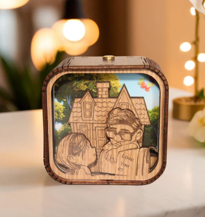 May include: A wooden music box featuring a laser-cut design of a couple and a house. The box has a square shape with a light brown frame and a detailed illustration inside. The words "Love You" are visible on a scroll. The background shows a blue sky and green trees.