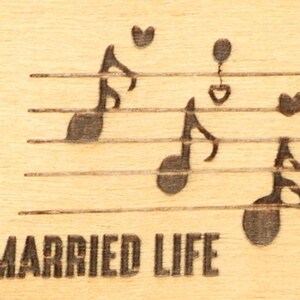 May include: A wooden plaque with engraved musical notes and hearts on a staff. The words "MARRIED LIFE" are etched below the staff. The background is a light wood grain.