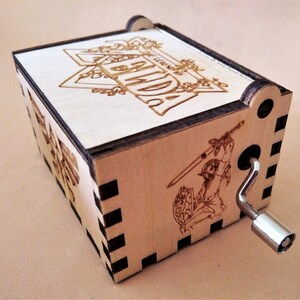 May include: A wooden music box with laser-etched designs. The top features the word "LEGEND" in a decorative frame. The side shows a warrior with a sword and shield. A silver crank is on the side. The box is made of light-colored wood.