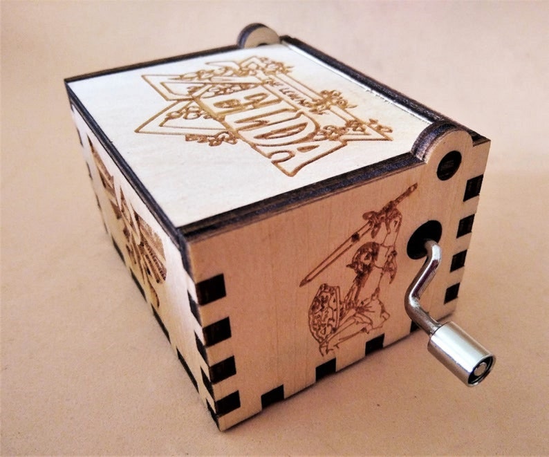 May include: A wooden music box with laser-etched designs. The top features the word "LEGEND" in a decorative frame. The side shows a warrior with a sword and shield. A silver crank is on the side. The box is made of light-colored wood.
