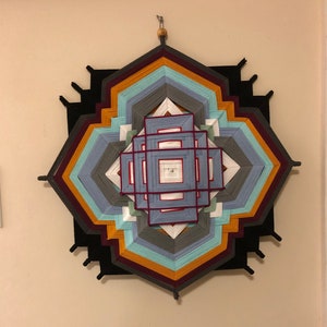 May include: A colorful geometric wall hanging with a central square design. The hanging is made of yarn in shades of red, orange, yellow, blue, green, and gray.