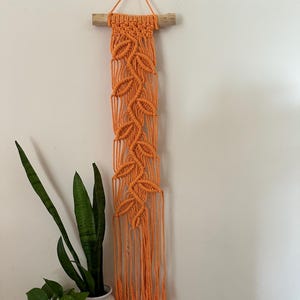 May include: Orange macrame wall hanging with a leaf pattern and fringe. The hanging is attached to a wooden dowel.