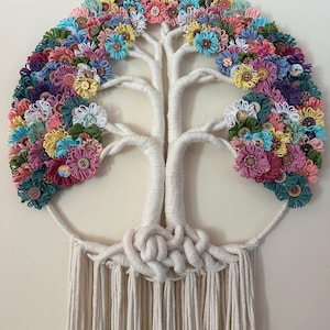 May include: A wall hanging featuring a white yarn-wrapped tree structure with a canopy of colorful yarn flowers and assorted buttons. The base of the tree is formed by intricate white cord knots, with long, straight white fringe hanging below.