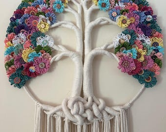 May include: A wall hanging featuring a white yarn-wrapped tree structure with a canopy of colorful yarn flowers and assorted buttons. The base of the tree is formed by intricate white cord knots, with long, straight white fringe hanging below.