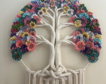 Op de afbeelding: Een wandhanger met een boomstructuur omwikkeld met gebroken wit garen, met een bladerdak bestaande uit talloze kleine, kleurrijke garenbloemen en knopen in tinten roze, blauw, geel, groen en paars, met lange franjes aan de onderkant.