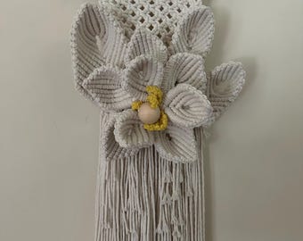 May include: A cream-colored macrame wall hanging featuring a large, textured flower with a yellow center and a wooden bead. The flower is the focal point, with detailed cord work and fringe. The piece is suspended from a natural wood dowel.