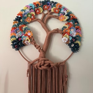 May include: A wall hanging featuring a tree silhouette wrapped in brown yarn, set within a circular frame. The branches are adorned with a dense arrangement of colorful fabric flowers and various buttons in shades of orange, blue, and yellow.