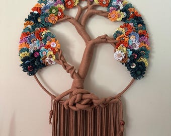 Op de afbeelding: Een wandhanger met een boom-silhouet omwikkeld met bruin garen, geplaatst in een cirkelvormig frame. De takken zijn versierd met een dichte opstelling van kleurrijke stoffen bloemen en diverse knopen in tinten van oranje, blauw en geel.