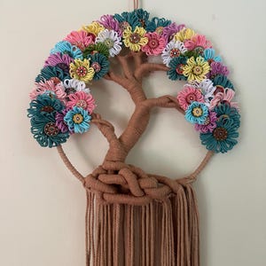 May include: A wall hanging featuring a brown yarn-wrapped tree structure with a circular frame. The branches are adorned with numerous small, colorful yarn flowers in shades of teal, pink, yellow, and white, each centered with a decorative button.