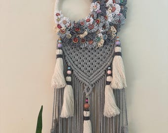 May include: A handmade macrame wall hanging featuring a crescent moon design adorned with fabric flowers and buttons in shades of gray, white, and red. The piece includes a gray macrame section with tassels and beaded accents, creating a bohemian aesthetic.