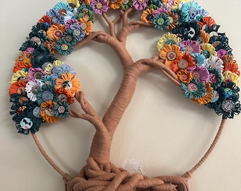 May include: A wall hanging featuring a tree structure wrapped in tan yarn, with a circular frame. The canopy is composed of numerous colorful yarn flowers and various patterned buttons in shades of orange, blue, yellow, and pink. Tan yarn fringe hangs from the base of the tree trunk.