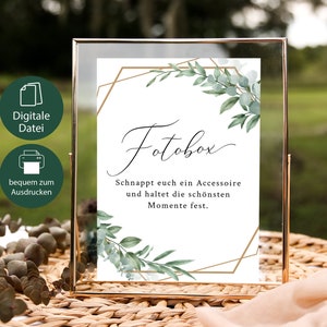 Op de afbeelding: Een afdrukbaar bord met een gouden geometrisch frame en groen blad, met de tekst "Fotobox" en "Schnappt euch ein Accessoire und haltet die schönsten Momente fest."