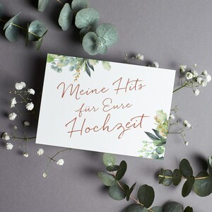 May include: A white card with green foliage border and the text "Meine Hits f&uuml;r Eure Hochzeit" in copper-colored lettering.