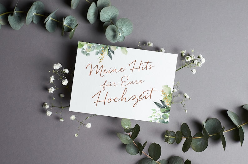 May include: A white card with green foliage border and the text "Meine Hits f&uuml;r Eure Hochzeit" in copper-colored lettering.