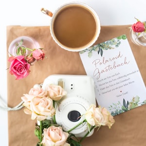 Peut inclure: Un appareil photo Polaroid blanc avec un livre d'or floral qui dit "Polaroid G&auml;stebuch" en texte brun. Le livre d'or contient des instructions pour que les invit&eacute;s prennent une photo, attendent qu'elle se d&eacute;veloppe, la collent dans le livre et laissent un message.