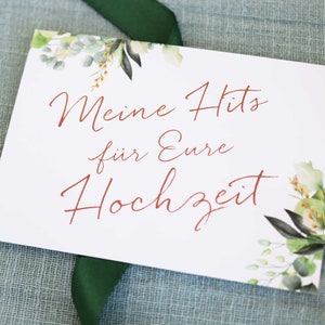 May include: A white card with green and brown floral illustrations on the top and bottom. The text "Meine Hits f&uuml;r Eure Hochzeit" is written in red cursive.