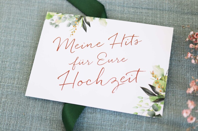 May include: A white card with green and brown floral illustrations on the top and bottom. The text "Meine Hits f&uuml;r Eure Hochzeit" is written in red cursive.