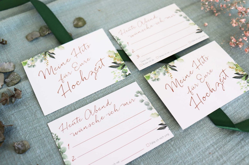 May include: Four white cards with green floral designs and copper calligraphy. The cards have the text "Meine Hits fur Eure Hochzeit" and "Heute Abend wunsche ich mir".