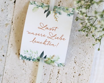 May include: A white card with green foliage and gold accents. The card has the text "Lasst unsere Liebe leuchten!" in copper lettering.