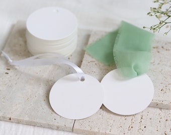 May include: A stack of white round gift tags with a hole punched in the center. The tags are on a light brown tiled surface with a white ribbon and a piece of light green fabric.
