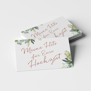 May include: A stack of white cards with green and gold floral designs. The text on the cards reads "Meine Hits f&uuml;r Eure Hochzeit".