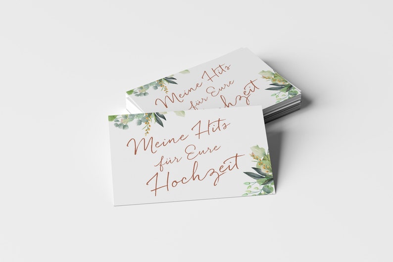 May include: A stack of white cards with green and gold floral designs. The text on the cards reads "Meine Hits f&uuml;r Eure Hochzeit".