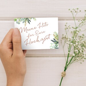 May include: A white card with green foliage and gold accents. The card says "Meine Hits f&uuml;r Eure Hochzeit" in copper-colored lettering.