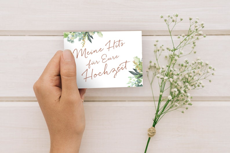 May include: A white card with green foliage and gold accents. The card says "Meine Hits f&uuml;r Eure Hochzeit" in copper-colored lettering.