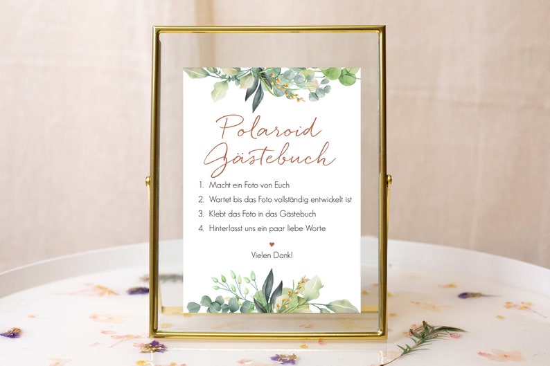 Peut inclure: Une pancarte blanche avec une bordure de feuillage vert dans un cadre dor&eacute;. La pancarte indique "Polaroid G&auml;stebuch" et contient des instructions pour un livre d'or Polaroid : 1. Prenez une photo de vous-m&ecirc;mes, 2. Attendez que la photo soit enti&egrave;rement d&eacute;velopp&eacute;e, 3. Collez la photo dans le livre d'or, 4. Laissez-nous quelques mots d'amour.  Vielen Dank!"