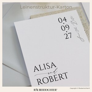 May include: A white card with black text that says "Leinenstruktur-Karton" at the top. The text "04.09.27" is centered on the card, with the text "Save the Date" written in cursive below it. The text "ALISA and ROBERT" is centered at the bottom of the card, with the text "R&Auml;UBERTOCHTER&reg;" below it. The text "Copyright &copy; R&auml;ubertochter&reg;" is in the bottom right corner of the card.