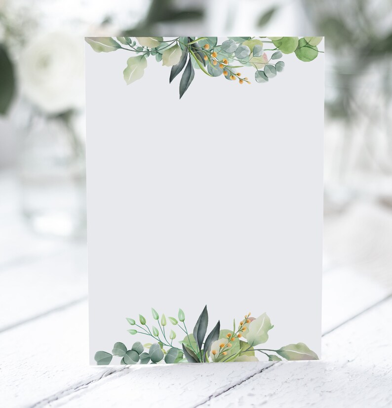 K&ouml;nnte beinhalten: Eine leere wei&szlig;e Karte mit einem gr&uuml;nen und gelben floralen Design an den oberen und unteren Ecken. Die Karte liegt auf einer wei&szlig;en Holzoberfl&auml;che.