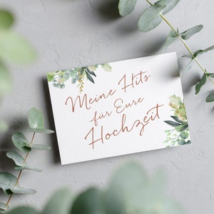 May include: A white card with green foliage on the edges. The text on the card reads "Meine Hits f&uuml;r Eure Hochzeit" in a copper-colored font.
