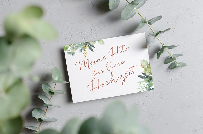 May include: A white card with green foliage on the edges. The text on the card reads "Meine Hits f&uuml;r Eure Hochzeit" in a copper-colored font.